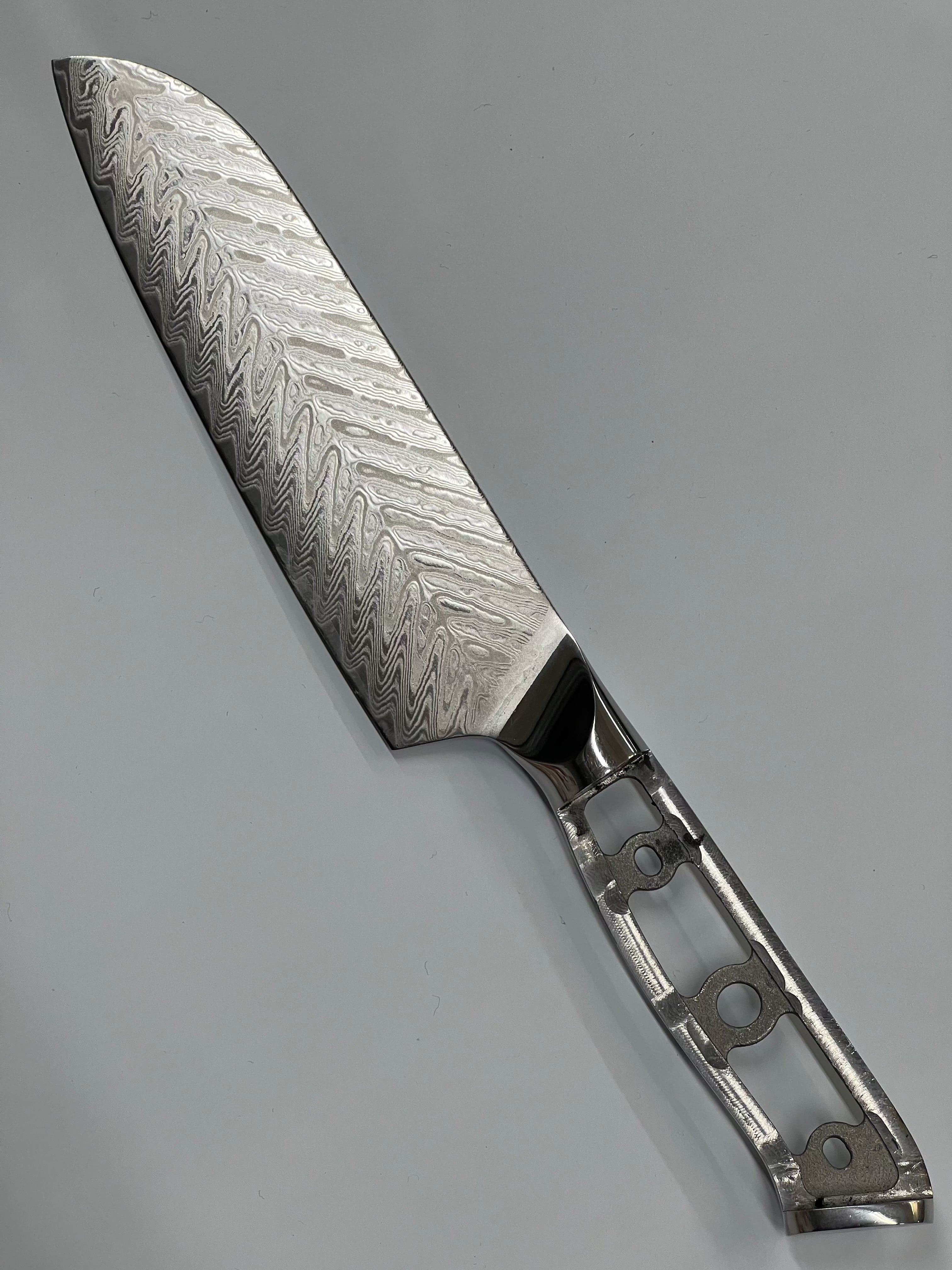 Et nærbilde av en Sild Santoku - VG10 Damask Kokkekniv 7" med et detaljert bølgemønster på bladet, laget med VG10 eggstål. Håndtaket er skjelettformet med sirkulære utskjæringer, noe som gir en moderne og lett design. Kniven hviler på en nøytral grå bakgrunn, og fremhever dets intrikate håndverk.